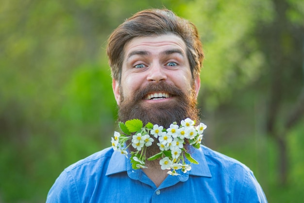 Un hombre barbudo con barba de flor divertida con vestido ligero mirando a la cámara mientras posa cerca de un árbol en flor en