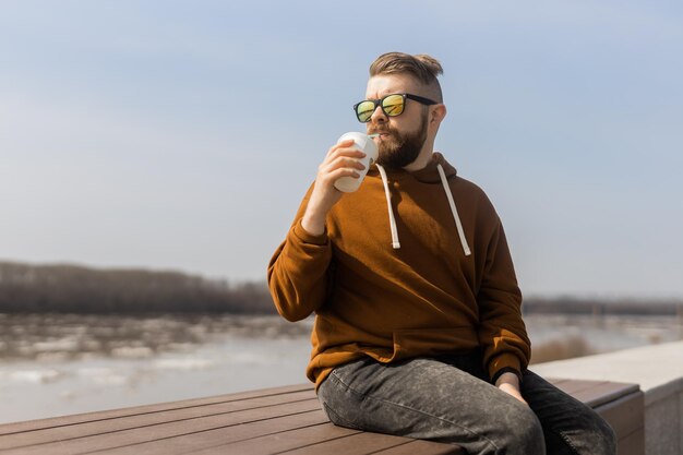 Hombre barbudo bebiendo un batido en el paseo marítimo de primavera bebidas para llevar y gen z o juventud milenaria