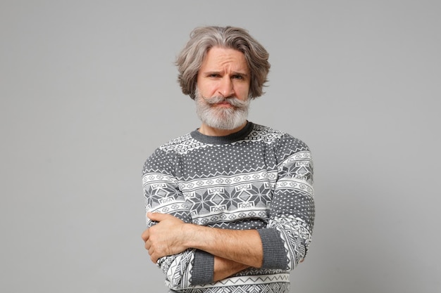 Hombre barbudo con bigote de pelo gris descontentado con suéter de punto posando aislado en el fondo de la pared gris, retrato de estudio. Concepto de estilo de vida de las personas. Simulacros de espacio de copia. Tomados de la mano cruzados.