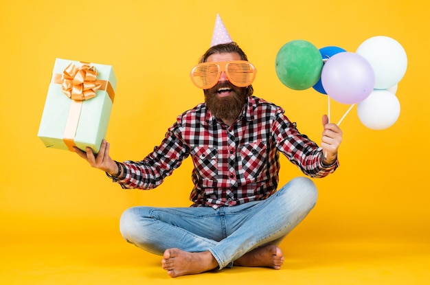 Un hombre barbudo brutal y divertido usa una camisa a cuadros con una barba y un bigote exuberantes, sostiene globos de fiesta y presenta una caja de cumpleaños