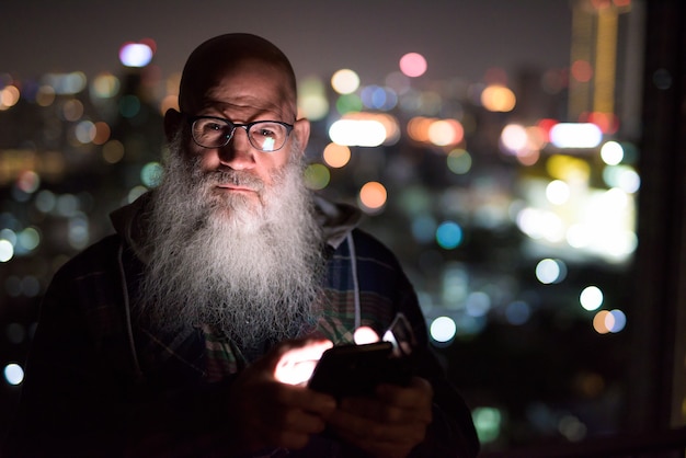 Hombre barbudo Calvo maduro con teléfono en vista de la ciudad por la noche