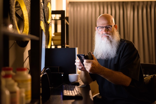 Hombre barbudo Calvo maduro usando el teléfono mientras trabaja horas extras en casa por la noche