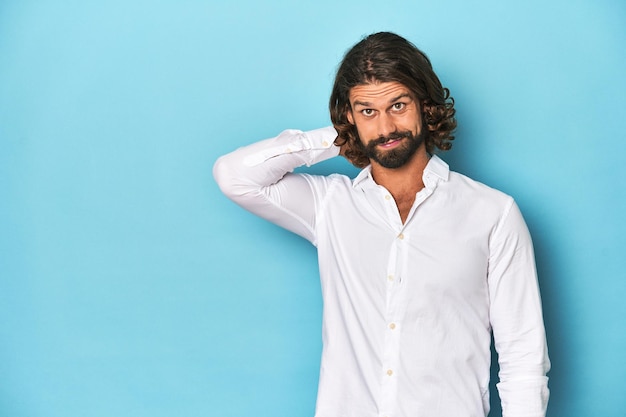 Hombre barbudo con una camisa blanca con telón de fondo azul tocando la parte trasera de la cabeza pensando y haciendo una elección