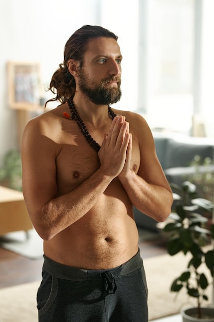 Hombre barbudo sin camisa de pie con las manos juntas mientras practica yoga en casa