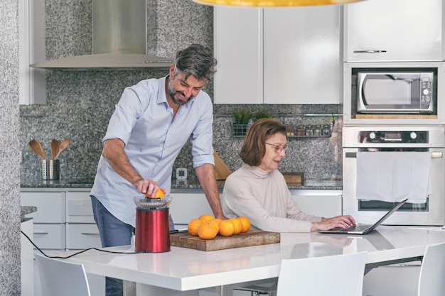 Hombre barbudo con camisa preparando jugo de naranja saludable en el exprimidor mientras una mujer mayor con gafas navega por la computadora portátil en la cocina en casa