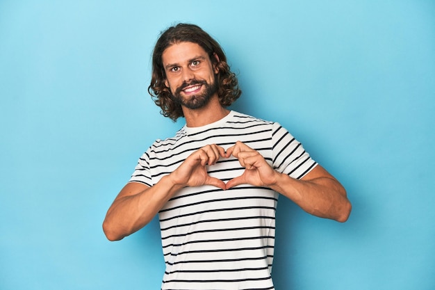 Hombre barbudo con una camisa a rayas con telón de fondo azul sonriendo y mostrando una forma de corazón con las manos