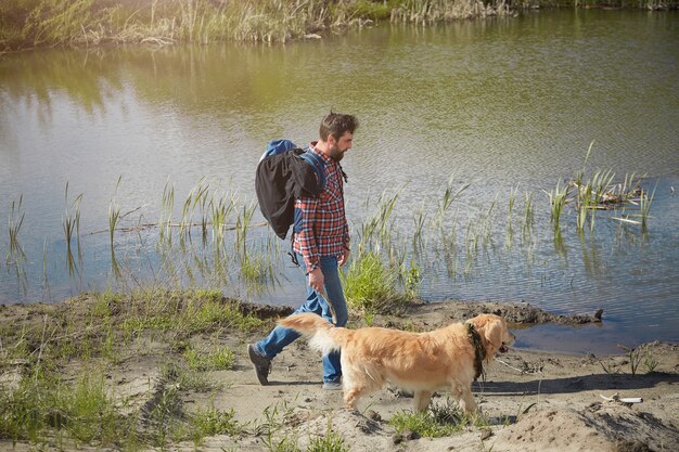 Un hombre barbudo con una camisa roja con un Golden Retriever atado, pasando por el lago.