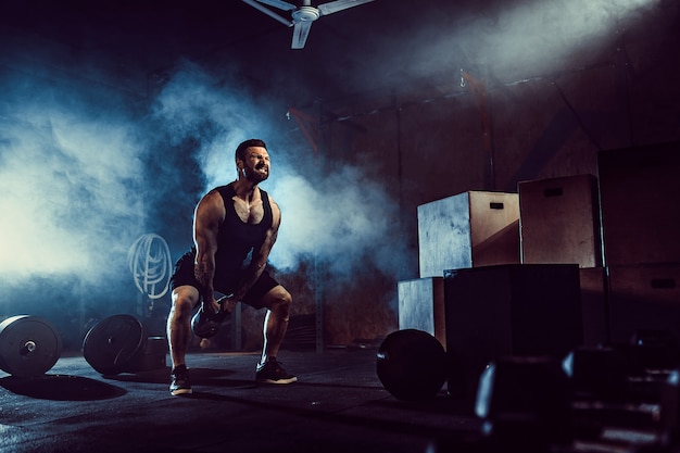 Hombre barbudo caucásico atractivo muscular que levanta dos pesas rusas en un gimnasio. Placas de pesas, mancuernas y neumáticos en el fondo.