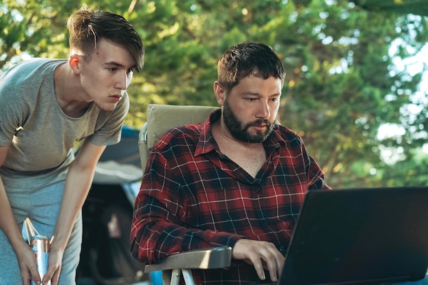 Foto hombre barbudo caucásico e hijo adolescente ala de trabajo con computadora portátil al aire libre concepto de camping