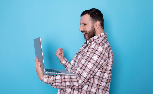Hombre barbudo caucásico feliz y emocionado sosteniendo una computadora portátil y levantando el brazo para celebrar el éxito