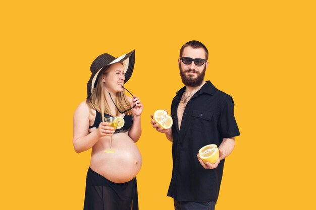 Un hombre barbudo y una chica embarazada en traje de baño sobre un fondo amarillo Frutas y bebidas refrescantes