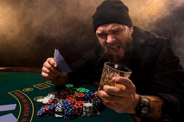 Hombre barbudo con cigarro y vidrio sentado en una mesa de póquer en un casino. Apuestas, naipes y ruleta.