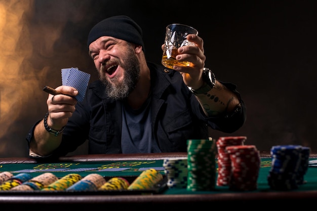 Hombre barbudo con cigarro y vidrio sentado en la mesa de póquer y gritando aislado en negro