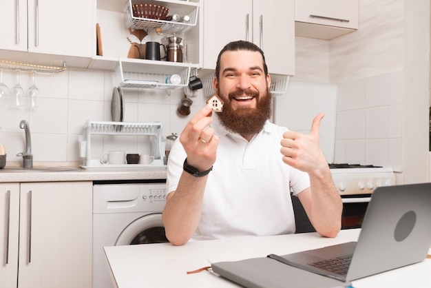 Hombre barbudo complacido sostiene una pequeña casa mientras está sentado en su cocina con su computadora portátil.