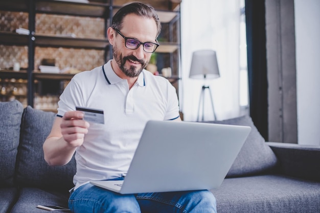 Hombre barbudo comprando en línea con tarjeta de crédito y usando una computadora portátil en casa