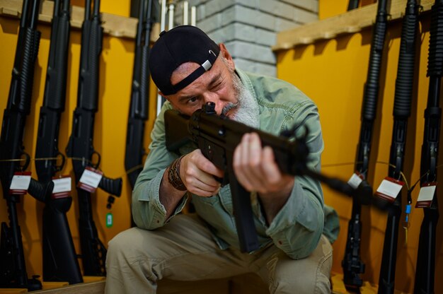 Hombre barbudo comprueba el alcance del rifle en la tienda de armas. Interior de la tienda de armas, surtido de municiones y municiones, elección de armas de fuego, pasatiempo y estilo de vida de tiro, autoprotección