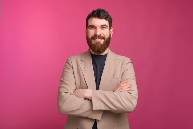 Hombre barbudo confiado con los brazos cruzados está sonriendo en la pared rosa