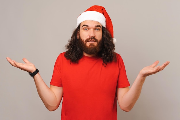 Hombre barbudo confundido se encoge de hombros mientras usa sombrero de Navidad en un estudio