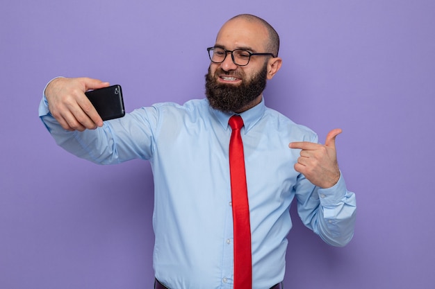 Hombre barbudo con corbata roja y camisa azul con gafas haciendo selfie con smartphone, sonriendo alegremente apuntando a sí mismo