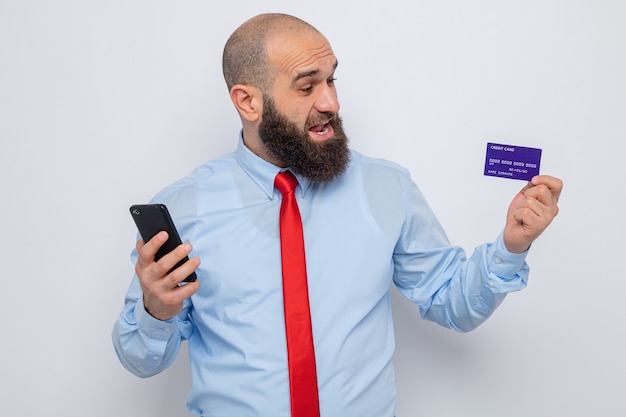 Hombre barbudo con corbata roja y camisa azul con smartphone y tarjeta de crédito mirando tarjeta sorprendido y feliz