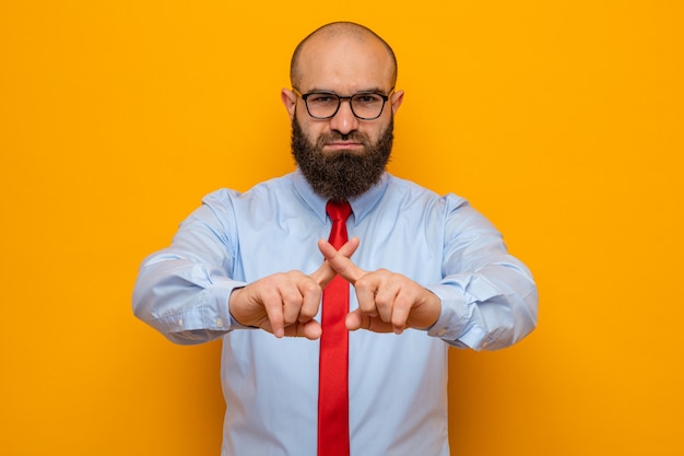 Foto hombre barbudo con corbata roja y camisa con gafas mirando a la cámara con cara seria haciendo gesto de parada cruzando los dedos índices de pie sobre fondo naranja