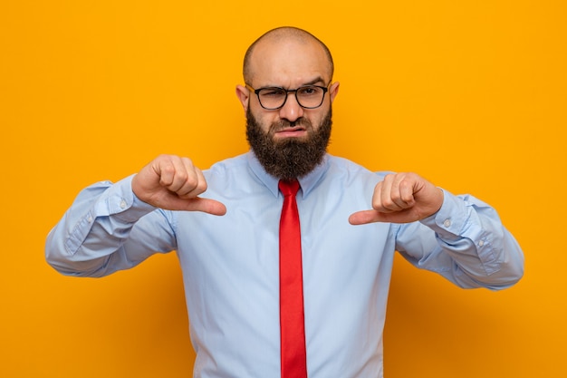 Hombre barbudo con corbata roja y camisa con gafas mirando con cara seria apuntando con los pulgares a sí mismo