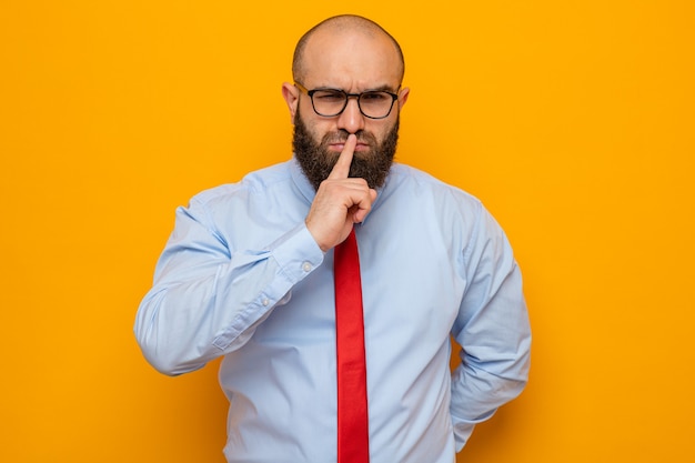 Hombre barbudo con corbata roja y camisa con gafas mirando con cara seria haciendo gesto de silencio con el dedo en los labios