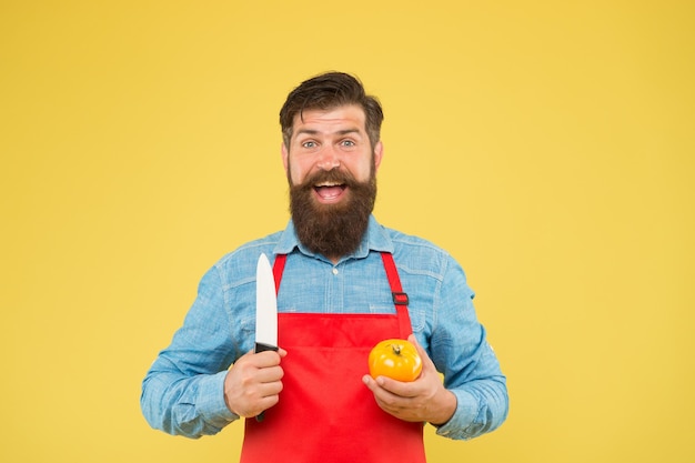 Hombre barbudo en delantal rojo con tomate amarillo cocinando alimentos saludables y frescos El chef masculino guapo está trabajando cocinero maduro cortado tipo vegetal en uniforme en la cocina dieta vitamínica