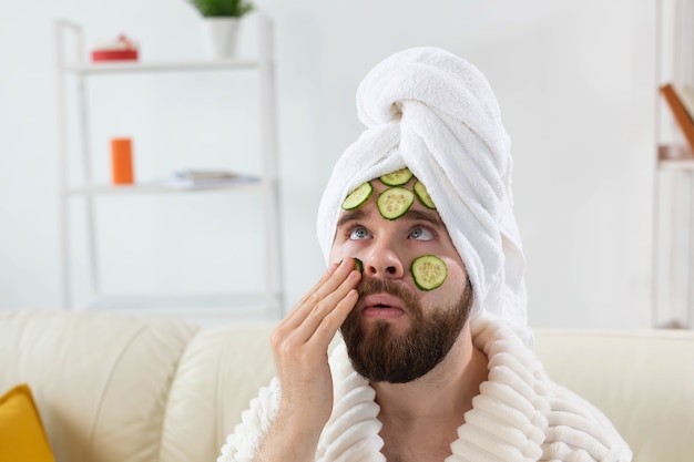 Hombre barbudo disfruta con una mascarilla cosmética en su rostro hecha de rodajas de pepino y cuidado de la piel de los hombres