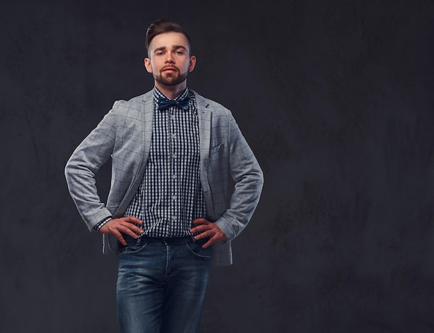 Hombre barbudo elegante y confiado con peinado en un elegante traje gris retro y pajarita, posando en un estudio. Aislado en un fondo gris.