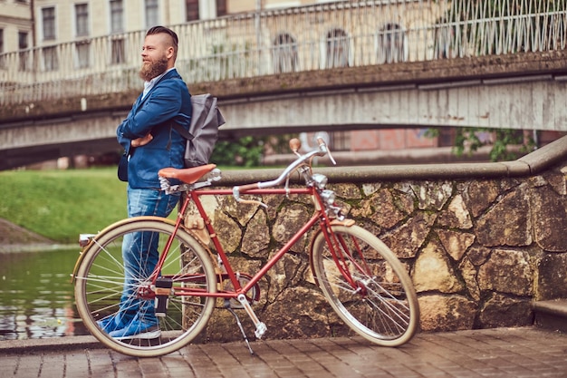 Un hombre barbudo con un elegante corte de pelo vestido con ropa informal con una mochila, de pie con una bicicleta retro cerca del río en un parque de la ciudad.