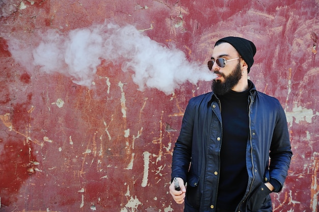 Hombre barbudo elegante joven en un sombrero negro con un cigarrillo electrónico o vape