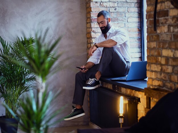 Hombre barbudo elegante trabaja con una computadora portátil en una habitación con interior de loft.