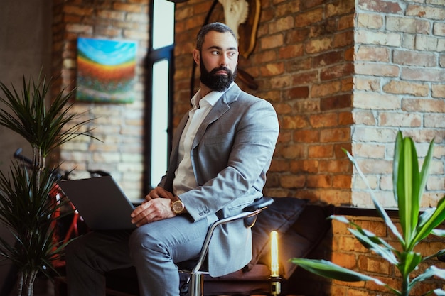 Hombre barbudo elegante trabaja con una computadora portátil en una habitación con interior de loft.