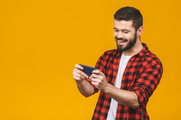 Hombre barbudo emocionado en camisa a cuadros jugando en smartphone aislado sobre la pared amarilla.