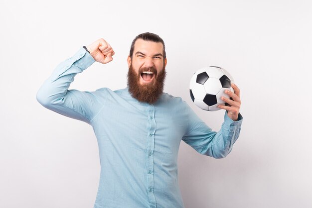 Hombre barbudo emocionado está haciendo el gesto ganador mientras grita y sostiene una pelota.