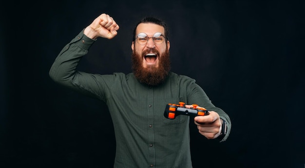 El hombre barbudo emocionado está haciendo el gesto ganador mientras sostiene un joystick Studio disparó sobre fondo negro
