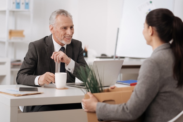 Hombre barbudo encantado positivo vestido con traje poniendo los brazos sobre la mesa manteniendo la taza en la mano derecha, mirando al nuevo colega