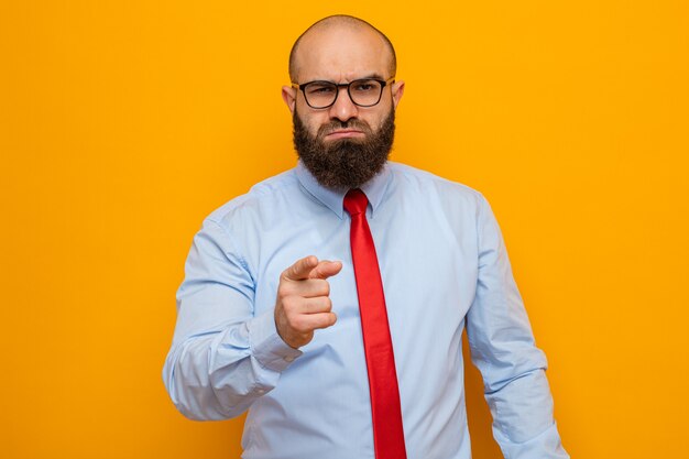 Hombre barbudo enojado con corbata roja y camisa con gafas apuntando con el dedo índice a la cámara con rostro serio de pie sobre fondo naranja