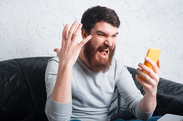 Hombre barbudo enojado gritando al teléfono inteligente mientras está sentado en el sofá en la oficina