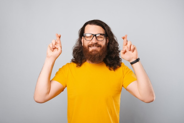 Un hombre barbudo esperanzado con una camiseta amarilla está de pie con los ojos cerrados y los dedos cruzados Foto de estudio sobre fondo gris