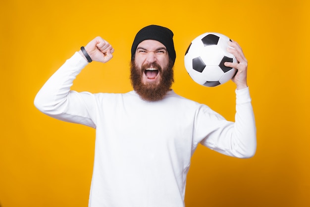 Un hombre barbudo está celebrando con una pelota de fútbol en la mano cerca de la pared amarilla.