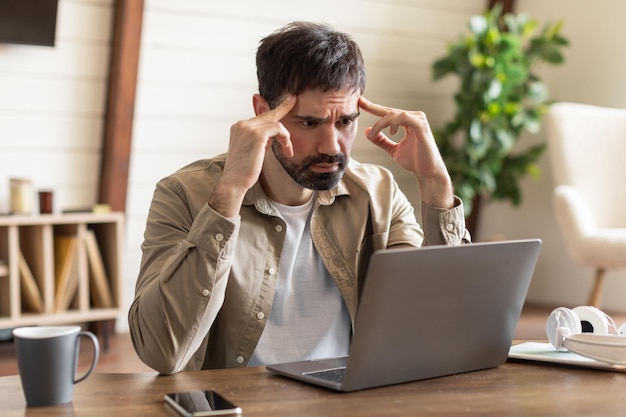 Un hombre barbudo está sentado en una mesa de madera con una computadora portátil que exuda una expresión de preocupación y profunda
