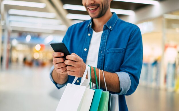 Hombre barbudo con estilo joven sonriente hermoso con bolsas de compras está usando el teléfono inteligente mientras camina en el centro comercial