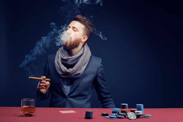 Hombre barbudo con estilo en traje y bufanda jugando en el casino oscuro fumar cigarro beber whisky