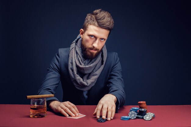 Hombre barbudo con estilo en traje y bufanda jugando en el casino oscuro fumar cigarro beber whisky