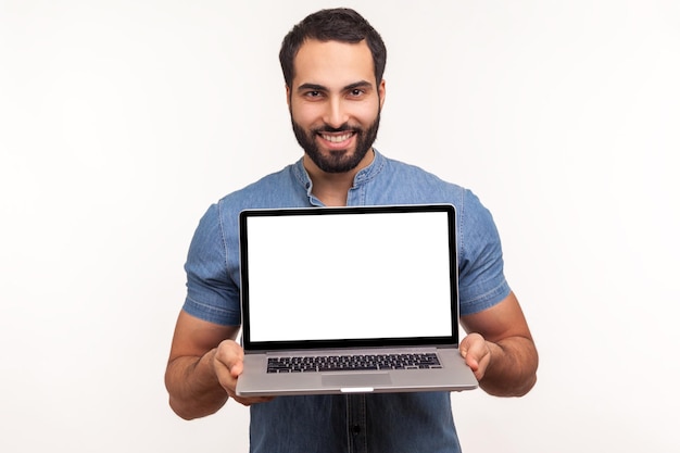 Hombre barbudo exitoso con camisa azul sosteniendo y mostrando una computadora portátil con una pantalla vacía en blanco, lugar para su anuncio, mirando la cámara, sonrisa con dientes. Disparo de estudio interior aislado sobre fondo blanco.