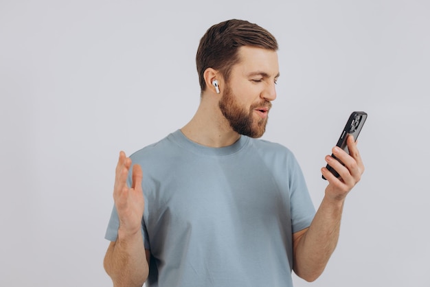 Hombre barbudo feliz bailando con auriculares escuchando música en auriculares con teléfono móvil y sonriendo complacido de pie sobre fondo blanco.