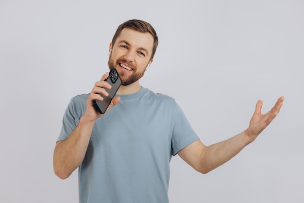 Hombre barbudo feliz bailando con auriculares escuchando música en auriculares con teléfono móvil y sonriendo complacido de pie sobre fondo blanco.