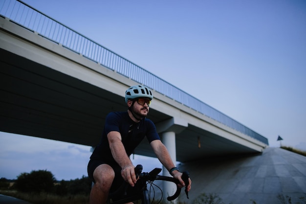 Hombre barbudo feliz con casco protector, gafas espejadas y ropa deportiva montando una bicicleta en una carretera asfaltada fuera de la ciudad por la noche